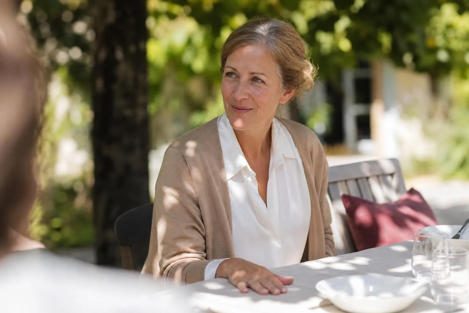 Femme d’une cinquantaine d’années assise à une table de jardin en fin de matinée, cheveux coiffés en chignon bas, pendant un repas de famille