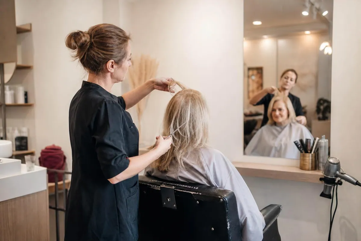Coiffeuse d’un salon français soulevant une mèche du dessus pour montrer une longueur de recouvrement à une cliente blonde d’une cinquantaine d’années assise au fauteuil, face au miroir